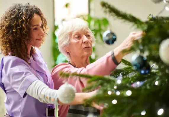 holiday home care - caregiver assisting senior woman add ornaments to christmas tree